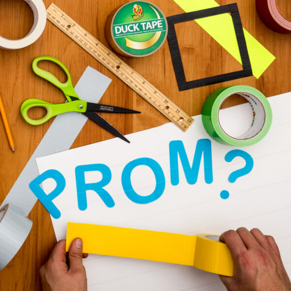 Hands laying down yellow duct tape on a poster board that says prom.