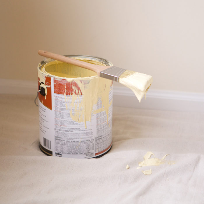 A bucket of white paint and a paint brush on a drop cloth.
