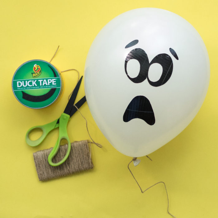 A balloon with a Duck tape ghost face on it, next to a pair of scissors and a roll of Duck Tape.