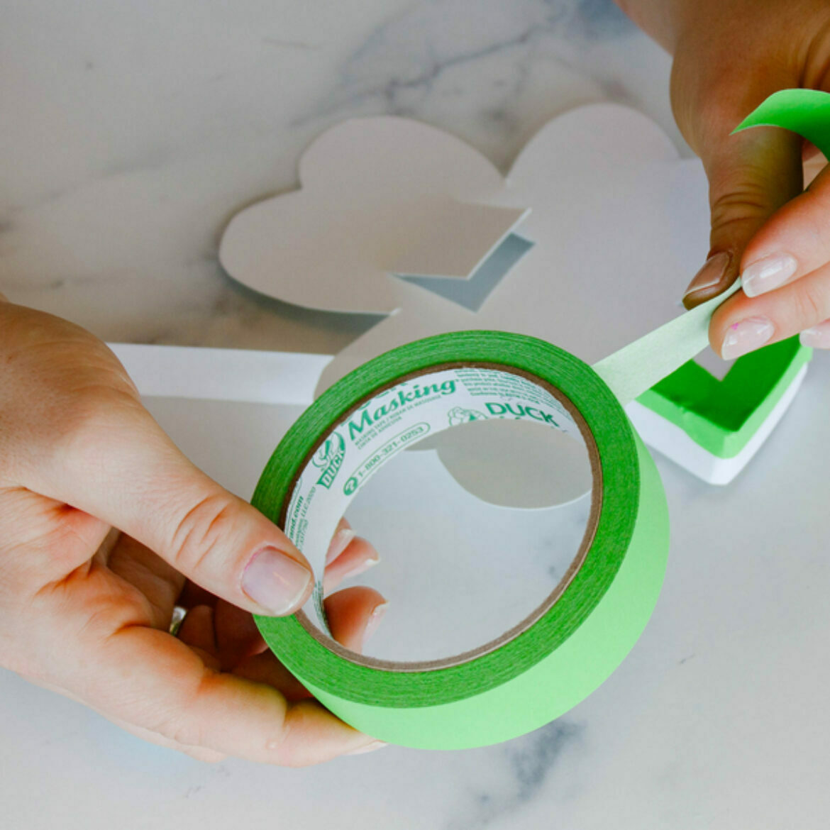 A white background with a white cut out of a shamrock and two hand holding a green roll of masking tape