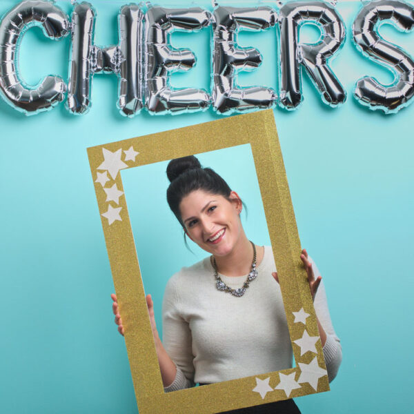 Woman posing with photo booth frame below "cheers" letter balloons